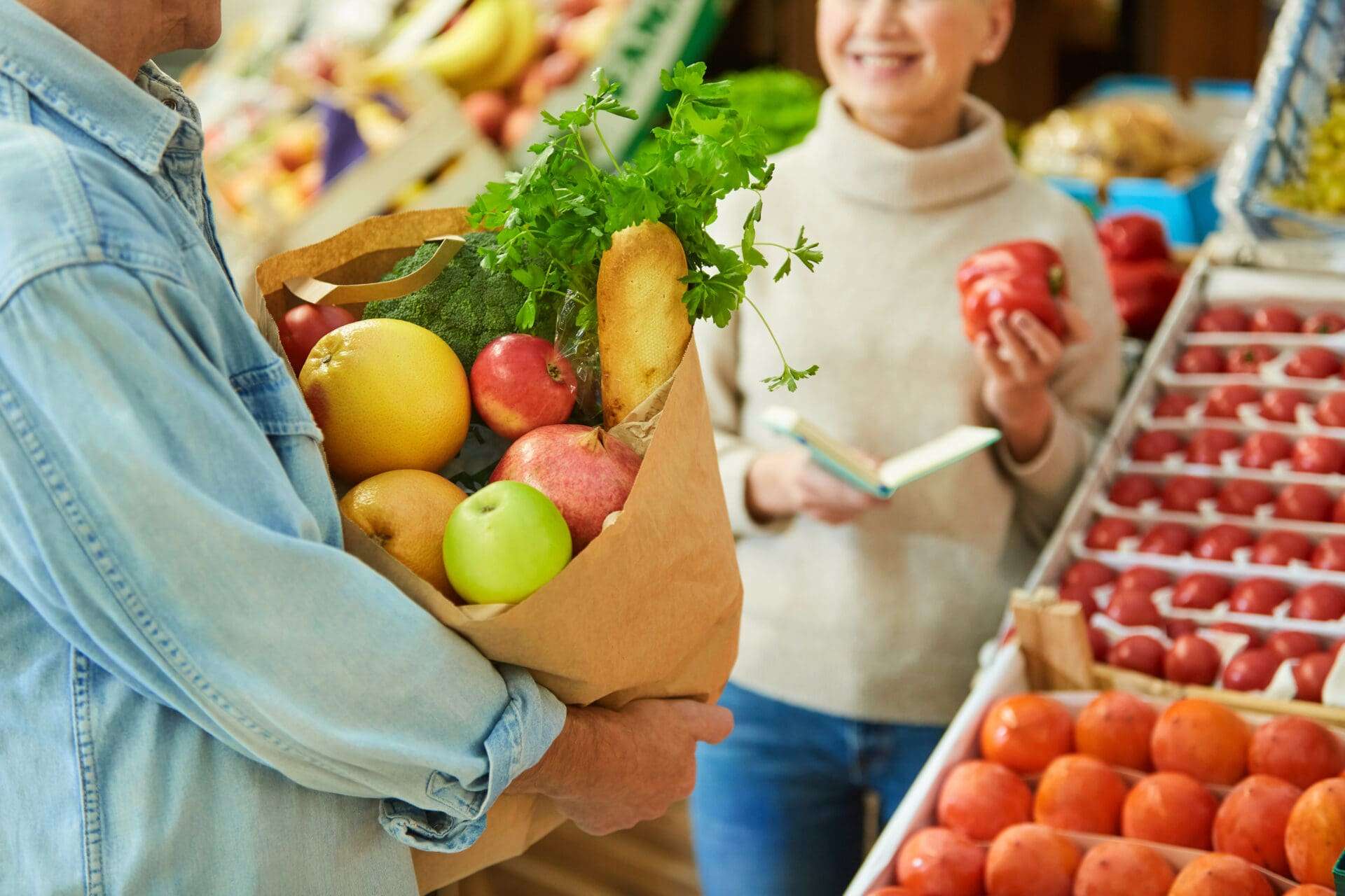 Close up of modern senior couple choosing fresh vegetables while enjoying shopping in farmers market, focus on paper bag with groceries in foreground